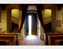 Innenraum eines historischen Gebäudes mit Holzbänken und rotbraun gefliestem Boden. Im Hintergrund ein großes Holztor mit rotem Vorhang, flankiert von zwei Treppen, die seitlich nach oben führen.