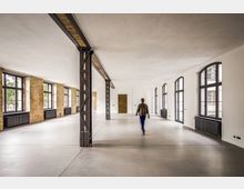 Großer, leerer Loft-Innenraum in einem umgebauten Industriegebäude mit freiliegenden Backsteinwänden, weißen Decken und Stahlstützen. Entlang beider Seiten verlaufen hohe, bogenförmige Fenster mit schwarzen Rahmen und darunterliegenden Heizkörpern über einem glatten Betonboden.