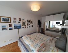 Schlafzimmer mit einem Doppelbett, farbigen Bettdecken mit Karomuster und Parkettboden. An der linken Wand hängen zahlreiche gerahmte Bilder, während rechts ein großer, weißer Kleiderschrank mit Spiegeltüren zu sehen ist; im Hintergrund fällt Tageslicht durch ein Fenster mit Vorhängen.