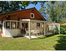 Ein kleines Haus mit Holz- und Putzfassade, ergänzt durch eine überdachte Terrasse mit weißen Gartenmöbeln. Der Garten ist von einer Rasenfläche und umliegenden Bäumen umgeben, die eine ruhige und grüne Umgebung schaffen.
