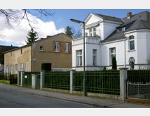 Straßenszene mit zwei Häusern: links ein schlicht verputztes Gebäude mit gelb-beige Fassade und rechts eine weiße Villa im historischen Stil mit Erker, Sprossenfenstern und Balkon. Ein Metallzaun mit Hecken grenzt die Grundstücke zur gepflasterten Straße ab, und eine Straßenlaterne steht am Gehweg.