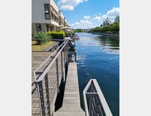 Mehrstöckige moderne Wohngebäude mit Terrassen am Ufer eines klaren Flusses, daneben ein schmaler Holzsteg und mehrere Boote. Im Hintergrund ist eine grüne Baumreihe sowie vereinzelt weitere Gebäude zu sehen, unter einem blauen Himmel mit einigen Wolken.