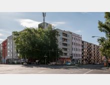 Straßenecke in einer städtischen Umgebung mit mehrstöckigen Wohngebäuden, Schaufenstern im Erdgeschoss und einem großen Baum im Vordergrund. Im Hintergrund ist eine kleine Kreuzung mit Ampeln und Verkehrsschildern zu sehen.