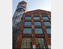 Backstein-Industriegebäude mit großen rechteckigen Fenstern und einem angrenzenden Turm mit Uhr. Die Architektur kombiniert rotbraune Backsteinfassaden mit Metall- und Glaselementen, unter einem klaren Himmel.