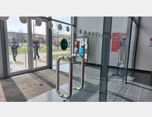 Blick aus einem modernen Eingangsbereich mit großen Glastüren auf einen Außenbereich mit Gehweg und Grünfläche. Im Inneren sind ein schwarzer Boden, ein schwarzer Fußabstreifer, ein Aushangbrett sowie ein Heizkörper sichtbar.