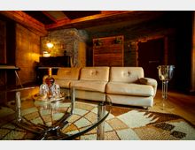 Wohnzimmer mit gemütlicher Einrichtung, bestehend aus einem cremefarbenen Sofa, einem Glastisch mit metallischen Beinen und einem gemusterten Teppich. Im Hintergrund ein Klavier, eine Tür und Holz- sowie Steinwandelemente, die den Raum warm gestalten.