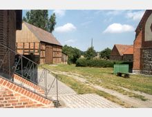 Ländliche Szenerie mit einer gepflasterten Fläche und Graswegen, umgeben von Gebäuden aus Ziegel- und Fachwerk. Im Vordergrund führen Treppen mit schmiedeeisernem Geländer zu einem Ziegelgebäude, im Hintergrund sind weitere Gebäude und Büsche sichtbar.