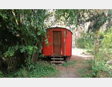 Ein roter Holzwagen mit Satteldach steht inmitten eines bewaldeten, naturbelassenen Gartens. Der Wagen hat eine kleine Holztreppe vor der Eingangstür und ist von Bäumen und Sträuchern umgeben.