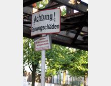 Ein Metallpfosten mit zwei Schildern unter einer rostigen Stahlbrücke; das obere Schild warnt vor Gehwegschäden, das untere vor Schnee und Glätte. Im Hintergrund sind grüne Bäume und ein Zaun zu sehen.