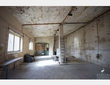 Großer, leerer Industrieraum mit abgenutzten Betonwänden und fleckiger Decke, an der Rohre und Leuchtstofflampen verlaufen. Links sorgen breite Fenster für Tageslicht; am Ende befindet sich eine grüne Doppeltür, daneben stehen eine Leiter und einige wenige Möbelstücke.