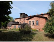 Backsteinbau mit Bogenfenstern und teils hölzernen Anbauten, umgeben von Bäumen und einer Wiese. Im Vordergrund steht eine kugelförmige Metallskulptur.
