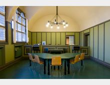 Raum mit grün gestrichenen Wandpaneelen und einem gewölbten Deckenbogen. In der Mitte steht ein grüner Tisch mit Stühlen aus Holz, während im Hintergrund eine Empfangstheke mit Durchgängen sichtbar ist; moderne Hängelampen und große Fenster mit Sprossen ergänzen das Design.