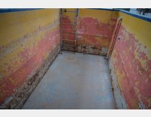 Ein kleiner, leerer, rechteckiger Behälter mit abblätternder gelber und roter Farbe an den Wänden, einer blauen Metallleiter an der Stirnseite und einem Ablassgitter in der Mitte des Bodens. Ein gelbes Rohr verläuft an der rechten Wand.