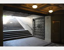 Unterführung mit einem Treppenaufgang, der nach draußen führt, und einer verzierten Metalltür an der rechten Wand. An der Decke sind mehrere runde Lampen angebracht, die den Bereich beleuchten.