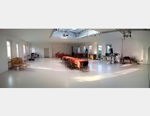 Heller, geräumiger Raum mit weißen Wänden und einem glänzend weißen Boden; in der Mitte steht ein langer Tisch mit orangener Tischdecke und Stühlen. Der Raum hat große Fenster an der Seite, eine Glasdecke mit Lichtinstallation und ist mit vereinzelten Möbelstücken sowie technischem Equipment ausgestattet.