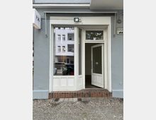 Fassade eines Gebäudes mit einer weißen Eingangstür und großen Fenstern, die teilweise beschriftet sind mit „F26 Pop-Up Store“. Der Gehweg besteht aus Kopfsteinpflaster, und im Fenster spiegelt sich ein gegenüberliegendes Gebäude.