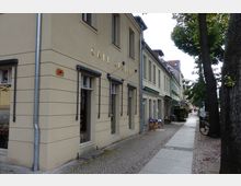 Straße mit Kopfsteinpflaster und angrenzenden beige gestrichenen Gebäuden, eines davon mit der goldenen Aufschrift "Café Claire". Im Vordergrund stehen Bäume, während entlang der Straße kleine Geschäfte und Fahrräder zu sehen sind.