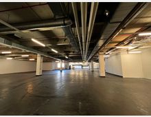 Weitläufiger leerer Industrie- oder Lagerraum mit Betonboden, mehreren weißen Stützsäulen und freiliegenden Rohren sowie Lüftungskanälen an der Decke. Die Wände sind glatt und in Weiß gestrichen, mit vereinzelter Beleuchtung entlang der Decke.