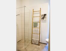 Modernes Badezimmer mit offener Dusche, gläserner Duschabtrennung, hellen Wand- und Bodenfliesen. An der Wand steht eine schmale Holzleiter mit aufgehängten Handtüchern, daneben hängt ein kleines Kunstbild.