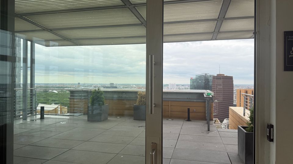 View through large glass doors onto a modern rooftop terrace with gray tiled flooring, metal railings, and square planters under a slatted canopy. A city skyline with several high-rise buildings is visible in the distance beyond the terrace edge.