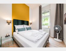 Modernes Schlafzimmer mit Doppelbett, hohem gepolstertem Kopfteil vor einer senfgelben Akzentwand und zwei Wandlampen. Großes Fenster mit grauen Vorhängen, hellem Holzfußboden sowie kleinem Glastisch und Standventilator neben dem Bett.