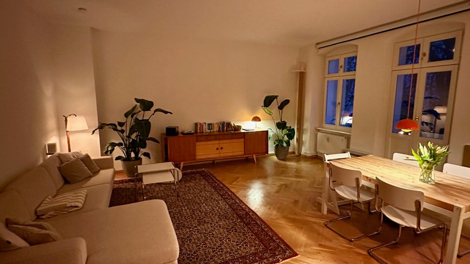 Gemütlicher Wohn- und Essbereich mit hellem Ecksofa, Teppich und Couchtisch sowie einem Esstisch mit Stühlen unter einer Hängelampe. Holzparkett im Fischgrätmuster, Sideboard mit Büchern, große Zimmerpflanzen und hohe Fenster mit Blick nach draußen.
