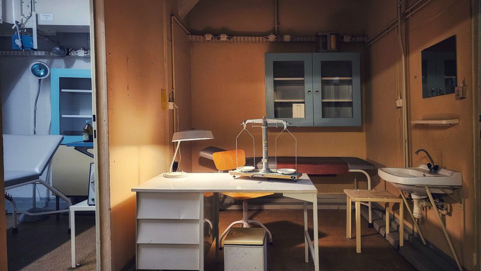 Innenraum eines alten Untersuchungs- oder Labors: ein Schreibtisch mit Lampe und Waage steht vor einer Liege, an der Wand hängen Glasschrank und Leitungen. Rechts befindet sich ein kleines Waschbecken, links ist durch eine Öffnung ein weiterer Raum mit Untersuchungsliege und Wandgerät zu sehen.