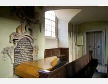 Innenraum einer historischen Kirche mit einer Gedenktafel an der Wand, die von einer Engelsfigur und Ornamenten flankiert wird. Im Vordergrund sind hölzerne Bänke und ein geschwungener Holzpult, im Hintergrund befindet sich eine Tür mit verzierten Paneelen.