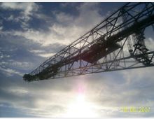 Ein großer Industriekran aus Metall erstreckt sich diagonal über den Himmel, silhouettiert von Sonnenlicht und umgeben von Wolken.