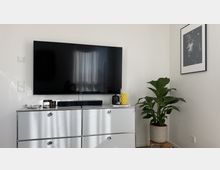 Moderne Wohnzimmerecke mit großem Flachbildfernseher auf einem silbernen Sideboard, darauf Soundbar und kleine Dekoobjekte. Rechts steht eine Zimmerpflanze im Topf neben einem gerahmten Schwarzweißfoto an der weißen Wand.