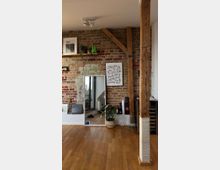 Wohnraum mit rustikaler Backsteinwand, sichtbaren Holzbalken und Holzfußboden. Im Vordergrund stehen Pflanzen, ein großer Spiegel lehnt an der Wand, daneben ein Regal mit dekorativen Gegenständen.