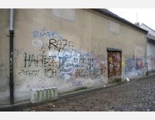 Eine Mauer an einer gepflasterten Straße, bedeckt mit farbigem Graffiti und Schriftzügen. Auf der rechten Seite befindet sich eine Holztür, und die Umgebung zeigt einen urbanen, etwas vernachlässigten Bereich.