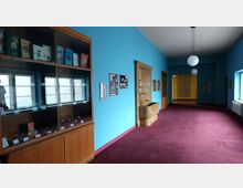 Ein Flur mit blauen Wänden und einem dunkelroten Teppichboden; links steht eine Holzvitrine mit Büchern und Exponaten, und rechts befindet sich ein dekorativer Wandbrunnen aus Stein. Große Fenster auf der linken Seite lassen Tageslicht ein, und runde Hängelampen sind an der Decke angebracht.