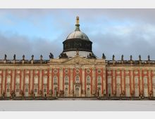 Barockschloss mit roter Fassade, verzierten weißen Fenstern und Statuen entlang des Dachrandes. Zentral befindet sich eine grüne Kuppel mit goldener Spitze; eine schneebedeckte Fläche liegt im Vordergrund.