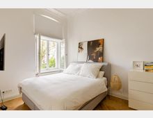 Helles Schlafzimmer mit einem Doppelbett, weißen Wänden und Holzboden. Ein großes Fenster mit Blick auf grünes Laub lässt Tageslicht herein, ergänzt durch moderne Dekoration wie ein abstraktes Gemälde und eine kleine Kommode.