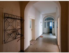 Langer, schmaler Flur in einem älteren Gebäude mit hohen Rundbögen, hellen Wänden und grauem Boden. Links hängt eine schwarze Metallgarderobe, und am Ende führen verglaste Doppeltüren unter einem Rundbogen in einen weiteren Raum.