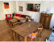 Wohnzimmer mit Holzfußboden, einem Holztisch, bunten Stühlen und einem Sofa mit gemusterten Kissen und Decke. Im Raum befinden sich ein roter Sessel, ein Wandspiegel, ein kleines Holzschränkchen und verschiedene Wanddekorationen.