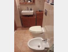 Ein Badezimmer mit beigem Fliesenboden und hellen Wandfliesen. Im Vordergrund ein WC und ein Bidet, im Hintergrund ein Waschbecken mit Holzunterschrank sowie ein kleines Regal mit Pflegeartikeln.