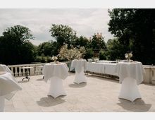 Steinerne Außenterrasse mit Balustrade, auf der mehrere Stehtische mit weißen Hussen und Blumenarrangements stehen; entlang der Seite steht ein langer Buffet- oder Getränketisch. Im Hintergrund ist ein grüner Park mit hohen Bäumen und ein entferntes schlossartiges Gebäude unter bewölktem Himmel zu sehen.