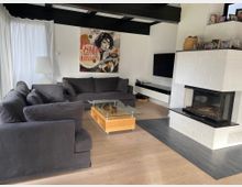 Wohnzimmer mit grauen Polstermöbeln, einem Glastisch auf einem Holzgestell und einer weißen gemauerten Kaminanlage. Im Hintergrund hängt ein farbenfrohes Kunstposter an einer weißen Wand, daneben ein flacher schwarzer Fernseher und ein modernes Sideboard.