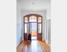 Helle Innenansicht eines Raums mit Holzboden und einer großen Holz-Doppeltür mit Glaseinsätzen, die zu einem Balkon mit ziegelroter Brüstung führt. Durch die Tür sind Gebäudedächer und ein wolkenloser Himmel sichtbar.