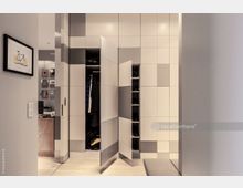 Ein moderner Eingangsbereich mit einem eingebauten Schrank im geometrischen Design aus weißen und grauen Paneelen; offene Türen geben den Blick auf Kleidung und Schuhe im Inneren frei. Im Hintergrund ist ein Schreibtisch mit Pinnwand zu erkennen, an der mehrere Gegenstände hängen.