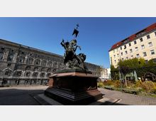 Bronzestatue eines Reiters, der einen Drachen bekämpft, auf einem großen steinernen Sockel in einem gepflasterten Platz. Im Hintergrund ein historisches Gebäude mit vielen Fenstern und ein begrünter Bereich neben einem gelben Wohnhaus.