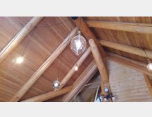 Holzdecke einer Hütte oder eines Gebäudes mit sichtbaren, massiven Holzbalken. Mehrere hängende Glühbirnen und eine große runde Lampe sind an dünnen Kabeln von der Decke abgehängt, im Hintergrund ist ein Druckmessgerät zu sehen.
