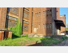 Backsteinfassade eines alten Industriegebäudes mit zahlreichen hohen Fenstern, teils vergittert, und einer Tür mit Treppenaufgang. Efeu wächst an einem Teil der Wand, und im Vordergrund befindet sich eine Grünfläche mit vereinzeltem Bewuchs.