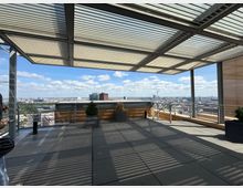 Rooftop terrace with large gray floor tiles, metal railings, and a slatted canopy that casts striped shadows across the deck. Planters line the edge, and the open side overlooks a wide city skyline under a partly cloudy sky.