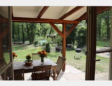 Ein überdachter Sitzbereich mit Holztisch und Stühlen, geschmückt mit Blumenvasen, führt in einen grünen Garten mit Bäumen. Im Garten stehen ein Grill, Gartenmöbel und Spielzeuge auf einer Rasenfläche.