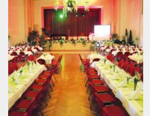 Ein festlich dekorierter Veranstaltungssaal mit Holzboden, langen Tischen mit weißen Tischdecken, roten Stühlen und grünen Tischdekorationen. Im Hintergrund gibt es eine Bühne mit geschlossenem roten Vorhang, dekorativem Pflanzenschmuck und einer Leinwand.