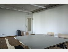 Ein moderner, minimalistisch eingerichteter Besprechungsraum mit weißen Wänden, einem rechteckigen Tisch mit Holzstuhlbestuhlung und einem Holzboden. Im Hintergrund befindet sich eine Tür mit Glaseinsatz und ein kleiner schwarzer Papierkorb.