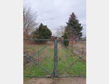 Ein verrostetes Metallgittertor mit Vorhängeschloss führt zu einem überwucherten Garten mit Gras und Bäumen. Im Hintergrund sind verzweigte Sträucher, ein Nadelbaum und eine kleine, verwitterte Hütte sichtbar, unter einem bewölkten Himmel.